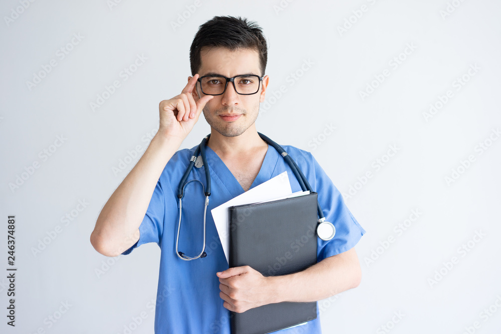 Serious young male doctor holding folder and documents. Handsome guy standing, adjusting glasses and wearing blue medical uniform. Professional doctor concept. Isolated front view on white background. Serious young male doctor holding folder and documents. Handsome guy standing, adjusting glasses and wearing blue medical uniform. Professional doctor concept. Isolated front view on white background.