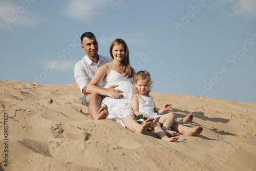 Happy family with daughter in white clothes are sitting toghether in desert sand dune.