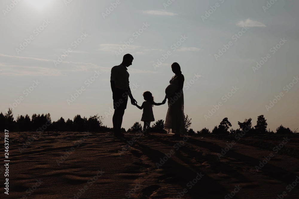 Silhouette portrait of beautiful young family in expectation of newborn. Man and woman standing face to face with their daughter over sunset sky. Background in evening in scenic nature landscape