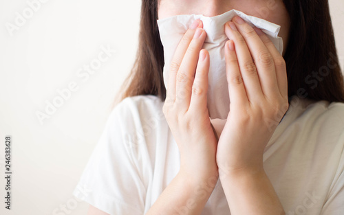 Young woman​ sneezing and blowing her nose w/ tissue​ paper. Cause of runny nose include common cold, flu, allergies, sinusitis, asthma, air pollution or influenza. Health care concept. Close up.