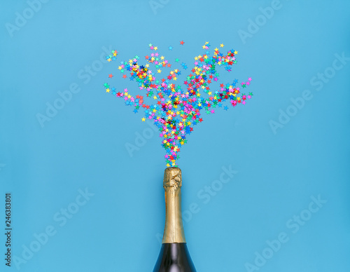 Creative photo of a champagne bottle with splashes of colorful confetti  on a blue background.Top view