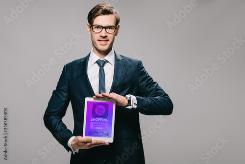 handsome businessman holding digital tablet with shopping app on screen isolated on grey