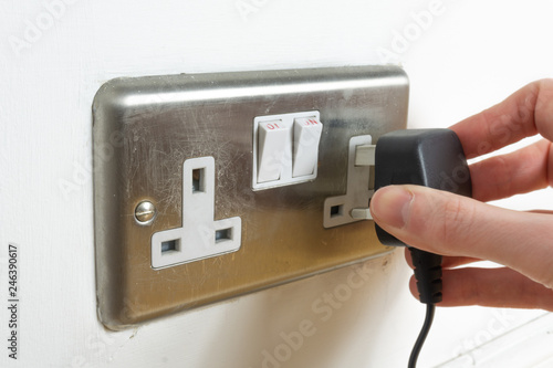 Stainless steel UK plug socket on white wall with hand plugging something in