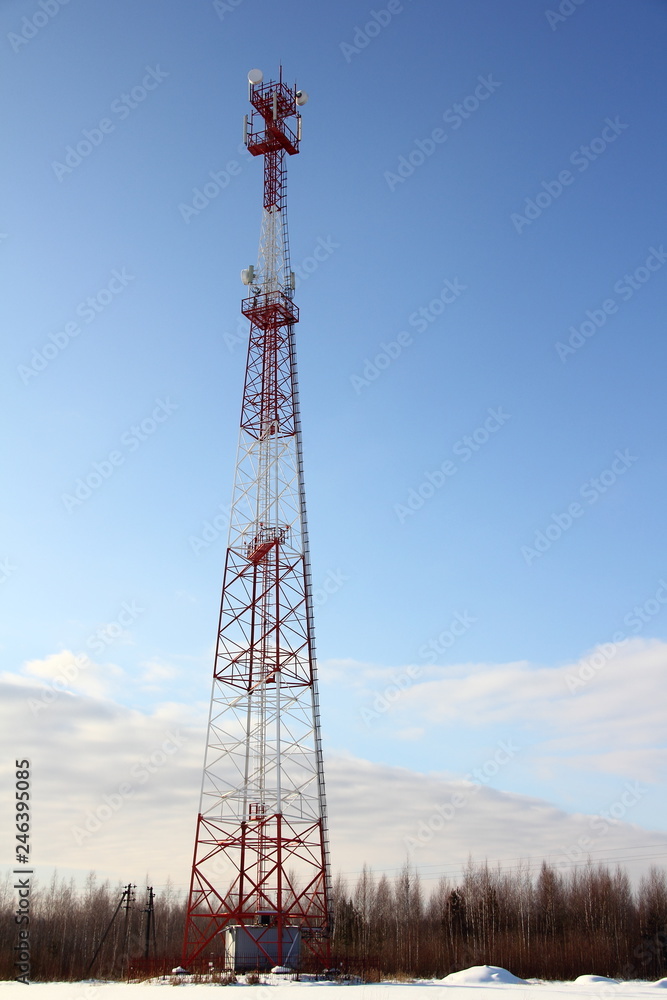 Big tower with a cellular antenna on a snowy field against the forest and blue sky on a winter day