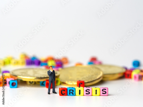 miniature figure businessman in dark blue suit standing backside of colorful of CRISIS alphabet and golden coin and thinking of the crisis will come.