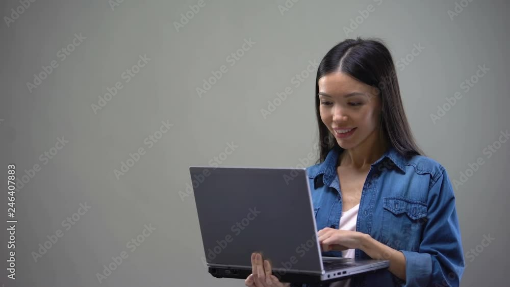 Cheerful asian woman typing on laptop standing on grey background, freelancer Cheerful asian woman typing on laptop standing on grey background, freelancer