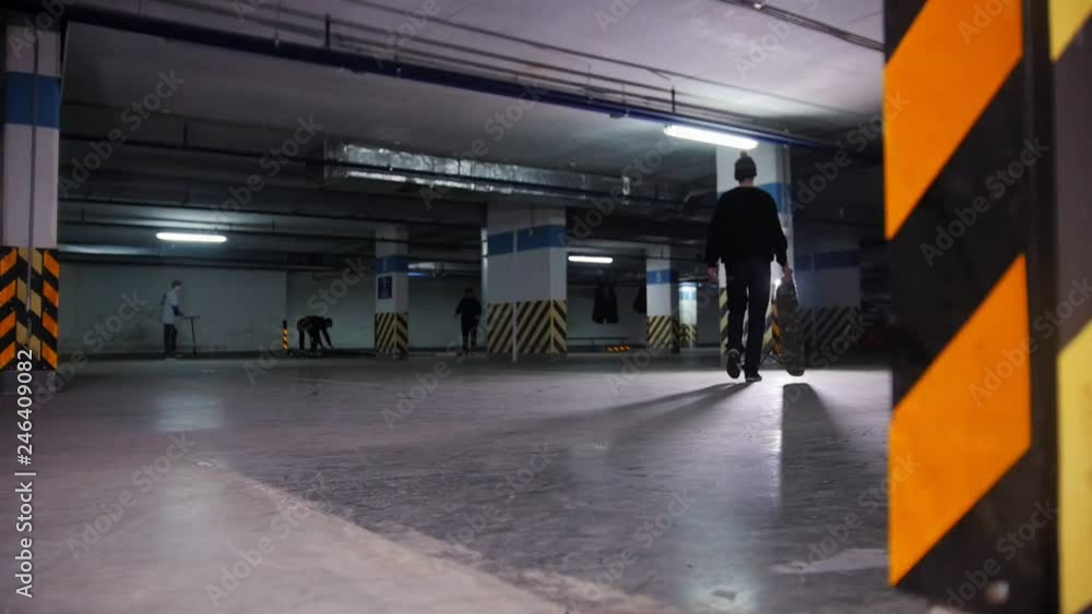 Skateboarding training. A young man skateboarding inside the parking lot, stops, takes the board in hand and goes towards the light. People training on the background Skateboarding training. A young man skateboarding inside the parking lot, stops, takes the board in hand and goes towards the light. People training on the background