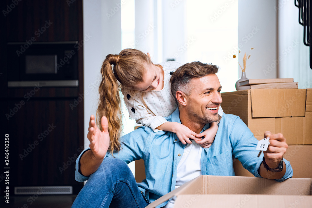 Fototapeta premium cheerful child hugging smiling father sitting on floor with house shaped key chain
