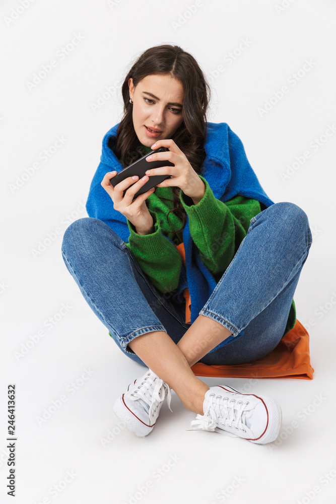 Image of cheerful woman 30s in colorful clothes sitting on the floor and using smartphone, isolated over white background