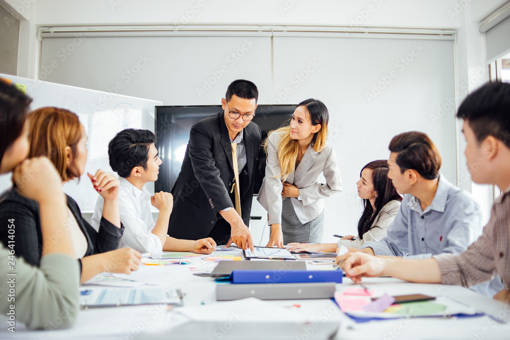 Businesswoman presenting to colleagues at a meeting.Successful team leader and business owner leading informal in-house business meeting. Businessman working on laptop in foreground. Businesswoman presenting to colleagues at a meeting.Successful team leader and business owner leading informal in-house business meeting. Businessman working on laptop in foreground.