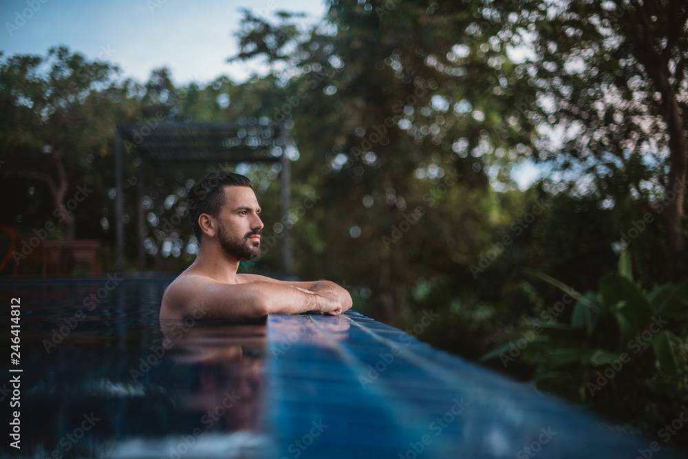 Fototapeta premium Portrait of a young handsome man in an outdoor pool in the tropics. Attractive guy with a beard at the side of the pool