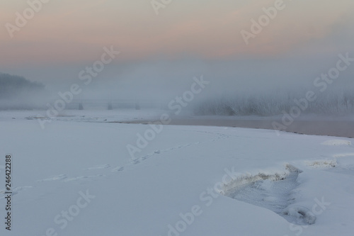 The river runs in the morning mist past the snowy shores and thickets of frost-covered bushes.