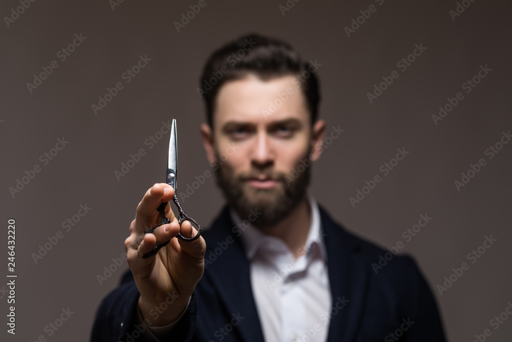 Young handsome bearded man with long beard moustache and brunette hair holding hairdresser or barber closed scissors with emotional face in studio on grey background Young handsome bearded man with long beard moustache and brunette hair holding hairdresser or barber closed scissors with emotional face in studio on grey background
