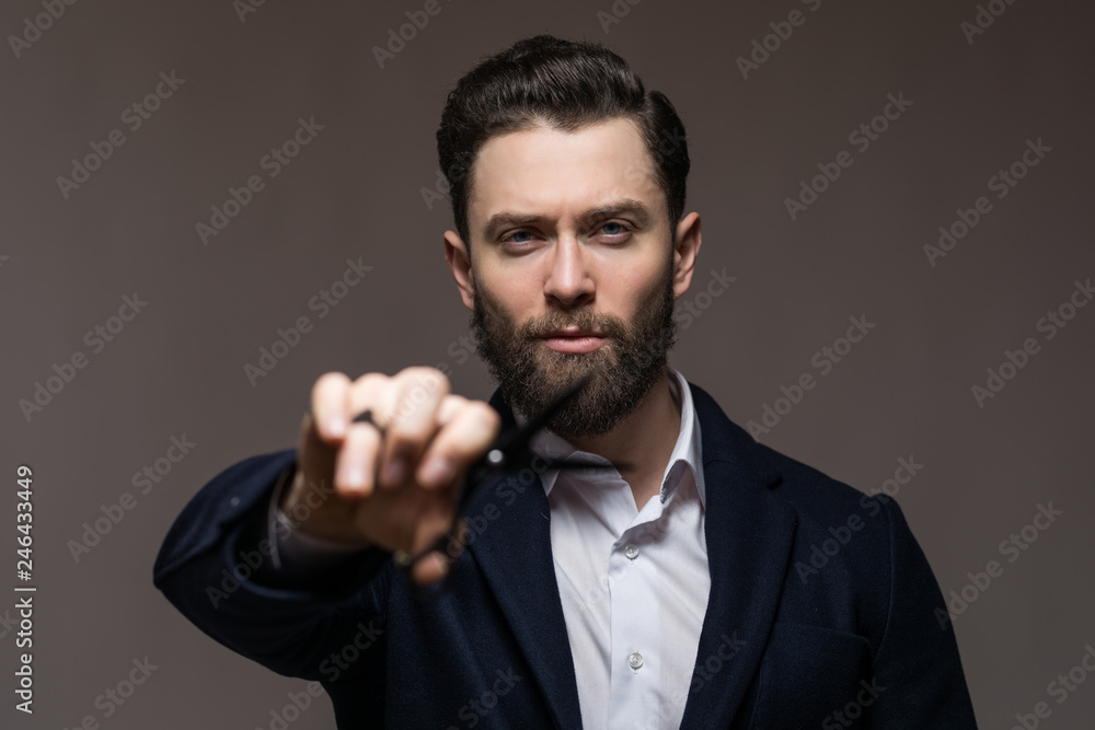 Young handsome bearded hipster man with long beard and brunette hair holding hairdresser or barber scissors and pointed it on camera with emotional face in studio on grey background. Focus on face. Young handsome bearded hipster man with long beard and brunette hair holding hairdresser or barber scissors and pointed it on camera with emotional face in studio on grey background. Focus on face.