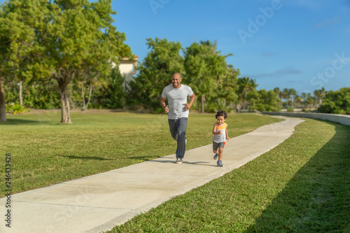 A father goes for a run with his little boy at the local park. The son keeps up with his dad during a morning workout on a sunny day.