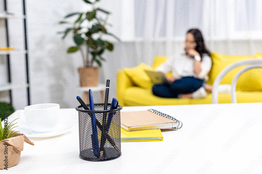 Selective focus of work desk with stationery with asian freelancer sitting on yellow sofa and using laptop on background Selective focus of work desk with stationery with asian freelancer sitting on yellow sofa and using laptop on background