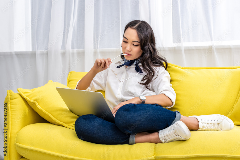 Young asian freelancer using laptop while sitting with feet on yellow sofa and holding glasses in hand Young asian freelancer using laptop while sitting with feet on yellow sofa and holding glasses in hand