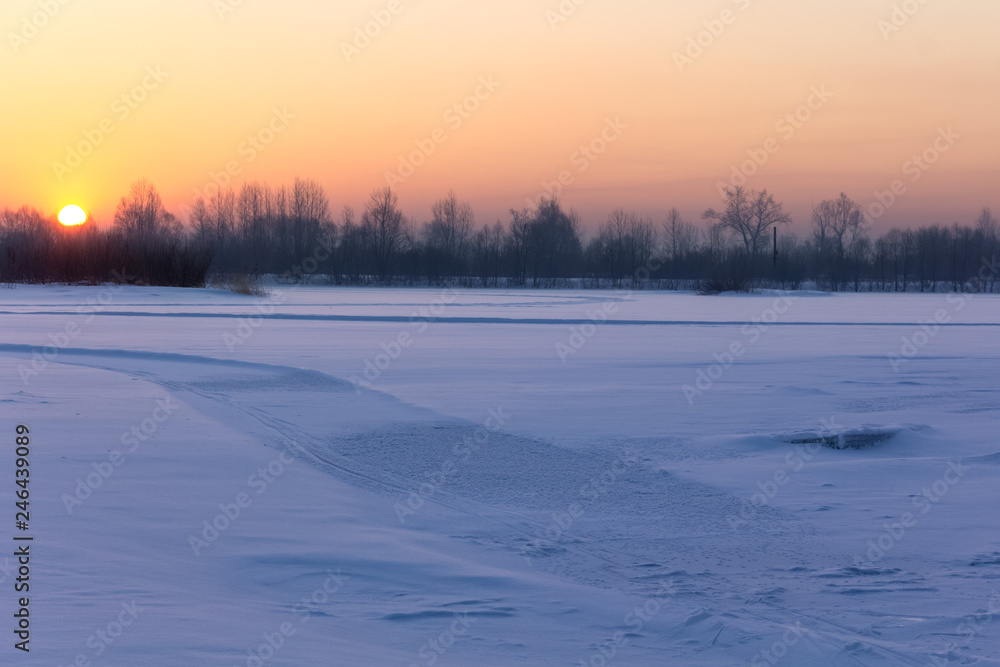 The bright sun rises from the horizon, illuminating the wide river covered with snow.
