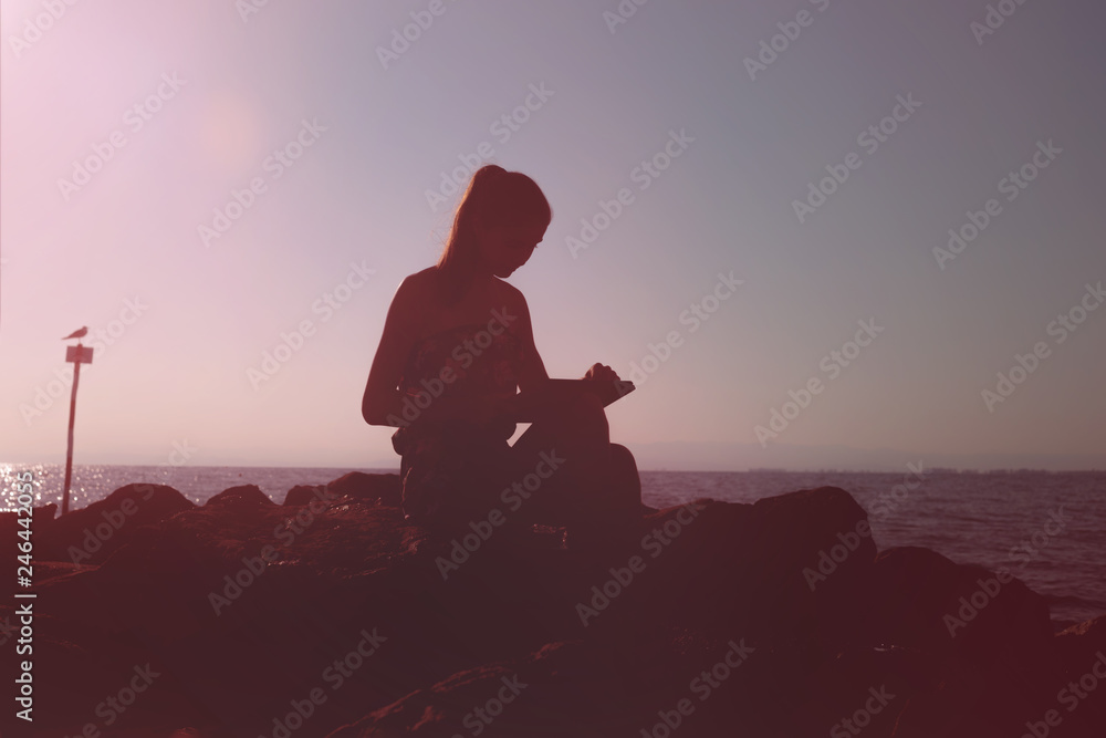 Silhouette of a girl sitting on the rocks near the sea, working on a laptop