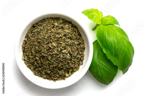 Dried chopped basil in white ceramic bowl next to fresh basil leaves isolated on white from above.