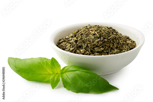 Dried chopped basil in white ceramic bowl next to fresh basil leaves isolated on white.