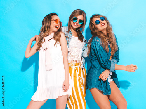Three young beautiful smiling hipster girls in trendy summer colorful clothes. Sexy carefree women in sunglasses isolated on blue. Positive models going crazy