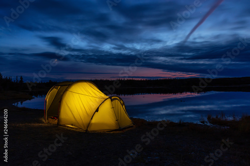 Lit Tent in nature near a calm lake during a vibrant sunset. Taken in Northern Newfoundland, Canada.