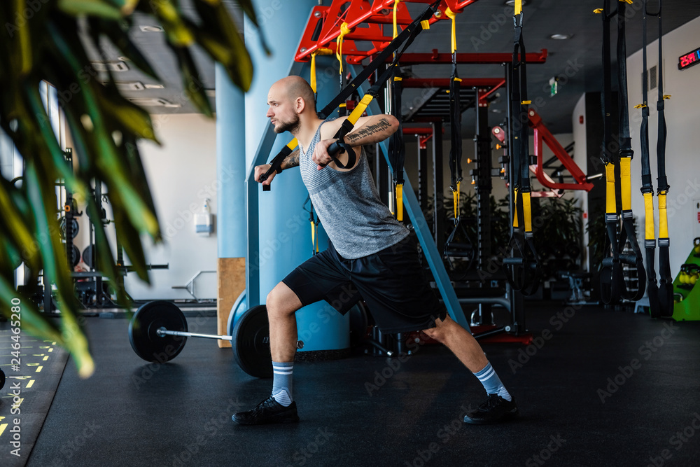 Φωτογραφία Tattooed athletic man exercising with suspension fitness rope in the gym