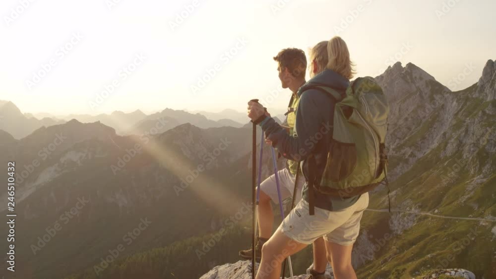 SLOW MOTION, LENS FLARE, CLOSE UP: Cheerful active tourists observe the stunning sunrise from a mountain peak. Happy Caucasian man and woman watch the sunrise high in the picturesque Slovenian Alps. SLOW MOTION, LENS FLARE, CLOSE UP: Cheerful active tourists observe the stunning sunrise from a mountain peak. Happy Caucasian man and woman watch the sunrise high in the picturesque Slovenian Alps.