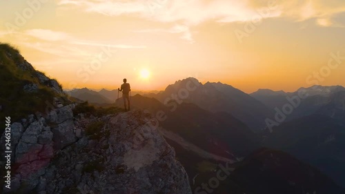AERIAL, SILHOUETTE, LENS FLARE: Stunning sunset illuminates the Alps and hiker standing on edge of a cliff and observing the picturesque mountain landscape. Man watching the sunrise in the mountains. photo