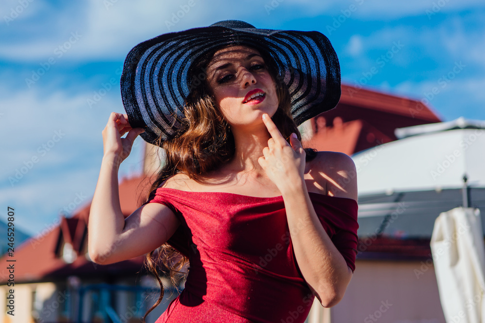 Beautiful young woman in red dress and balack hat in summer sunny day on the blue sky background.
