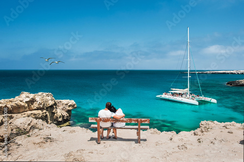 Honeymoon traveller couple hugging on a wooden bench and enjoys their tropical holiday. Wedding travel. Young happy couple sitting back on beach. Rear view of a couple in love on vacation. Travelers