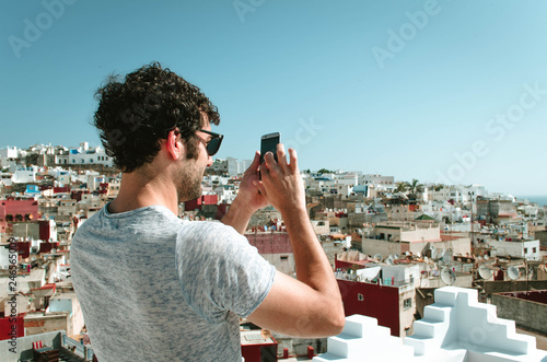 Portrait of happy young man on Morocco, tourist with smartphone taking pictures of old city Tangier from terrace