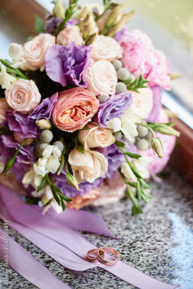 Close up of bridal bouquet of pink roses, peonies, purple flowers and herbs and two golden rings on marble window sill, copy space. Wedding concept. Selective focuse
