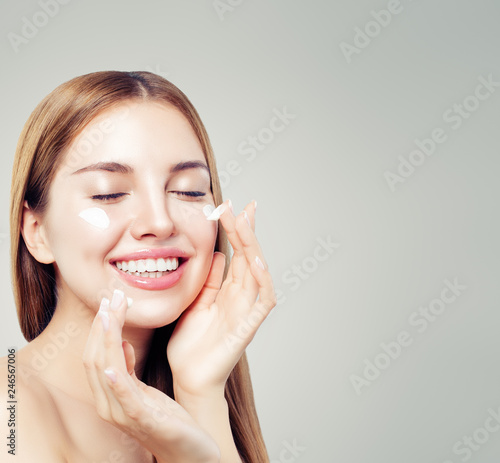 Laughing woman with healthy skin applying moisturizing cream on her face. Skin care, beauty and facial treatment concept
