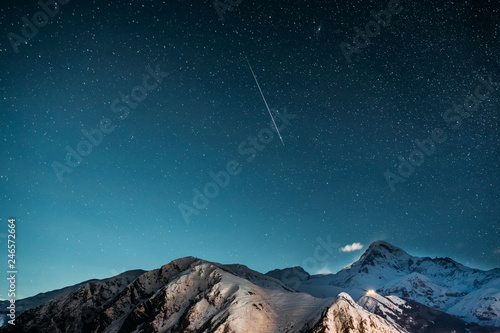 Stepantsminda, Georgia. Natural Winter Night Starry Sky With Glowing Stars And Real Meteoric Track Trail Over Mount Kazbek. Beautiful Night Georgian Landscape With Glowing Stars And Meteorite Trail