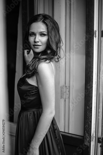 Black white portrait of young woman in elegant silk black dress lookingto a city from a balcony, fashion beauty photo