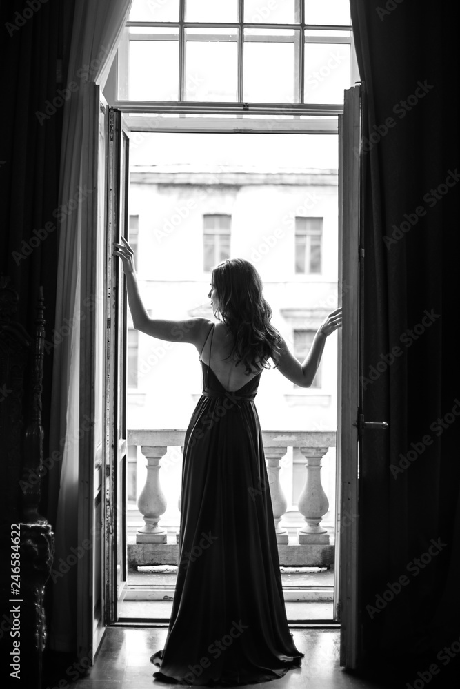 Fototapeta Czarno biały portret młodej kobiety w eleganckiej jedwabnej czarnej sukni, patrząca na miasto z balkonu.