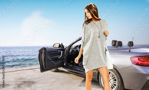 Slim young rich woman and sliver summer car on coast. Sea landscape and blue sky. 