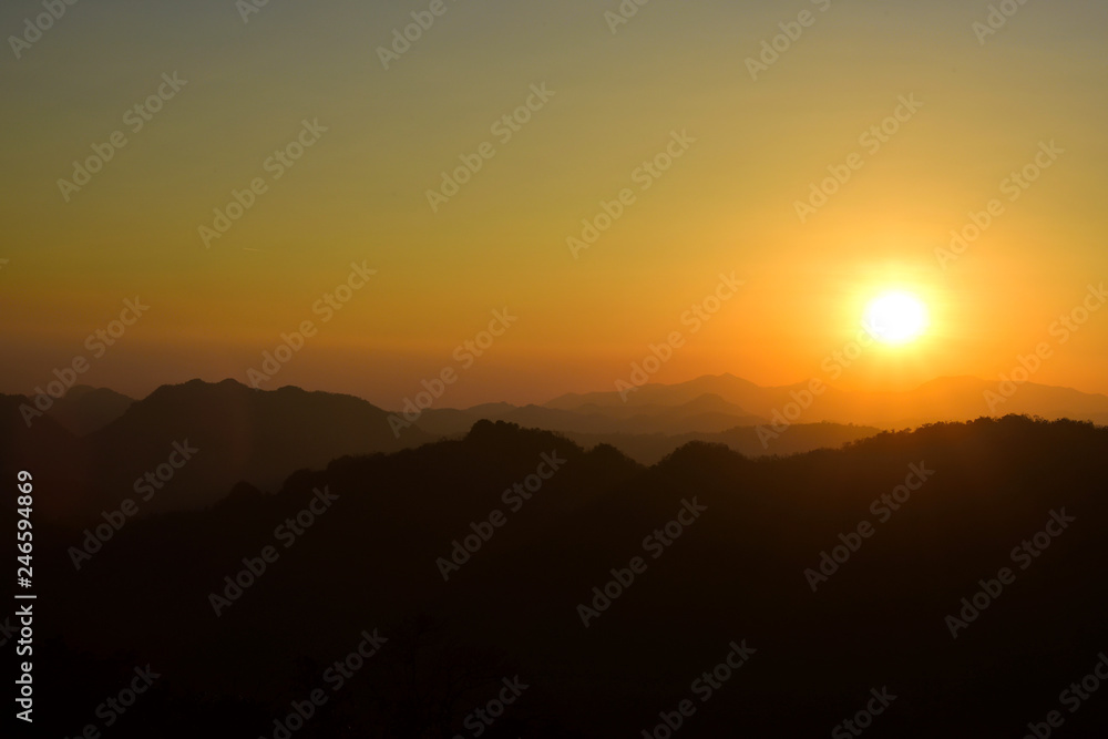 Picture of a sun setting behind a dense forest area followed by mountains.