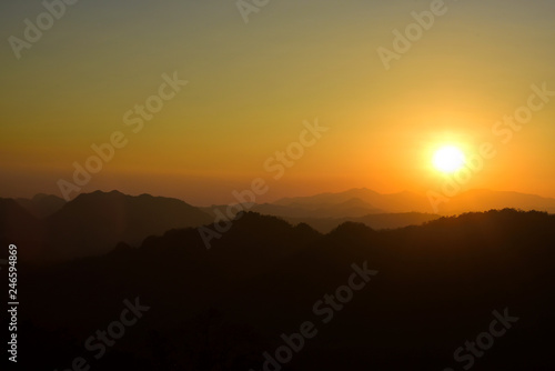 Picture of a sun setting behind a dense forest area followed by mountains. © tharathip
