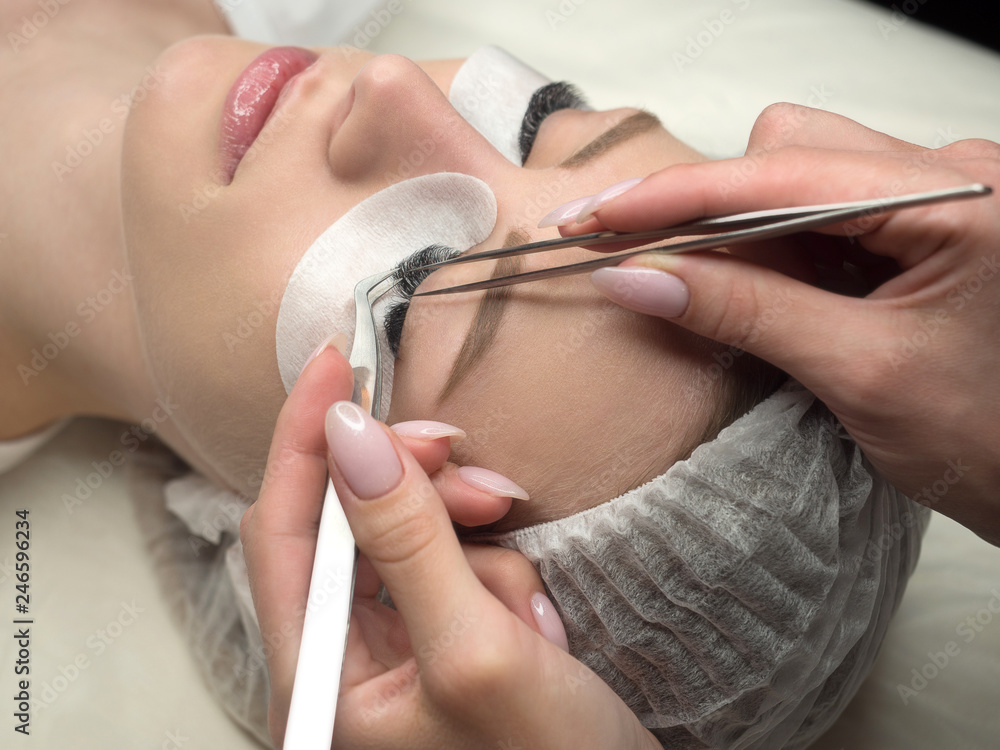 Eyelash extension procedure. Woman face and experts fingers with tweezers, close up, selective focus