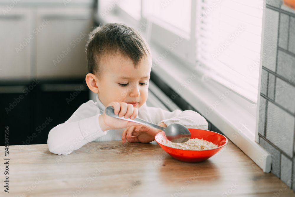 A child in the kitchen eating their own oatmeal with a red plate A child in the kitchen eating their own oatmeal with a red plate
