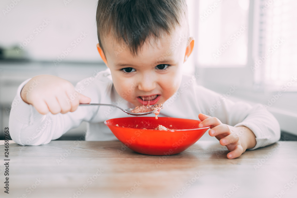 A child in the kitchen eating their own oatmeal with a red plate A child in the kitchen eating their own oatmeal with a red plate