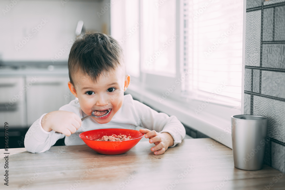 A child in the kitchen eating their own oatmeal with a red plate A child in the kitchen eating their own oatmeal with a red plate