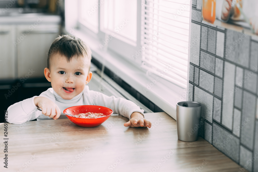 A child in the kitchen eating their own oatmeal with a red plate A child in the kitchen eating their own oatmeal with a red plate
