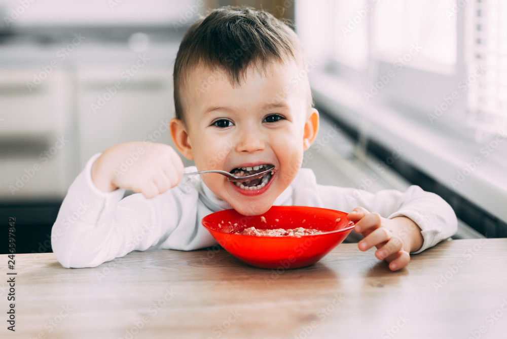 A child in the kitchen eating their own oatmeal with a red plate A child in the kitchen eating their own oatmeal with a red plate