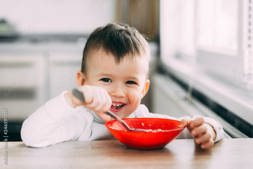 A child in the kitchen eating their own oatmeal with a red plate A child in the kitchen eating their own oatmeal with a red plate