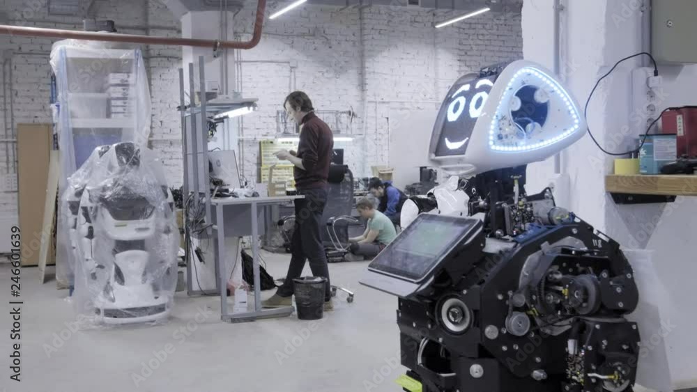 Factory for the production of robots, modern robotic developments. The disassembled robot waves his hands in greeting and dances in the workshop. Engineers walk in the background. Factory for the production of robots, modern robotic developments. The disassembled robot waves his hands in greeting and dances in the workshop. Engineers walk in the background.