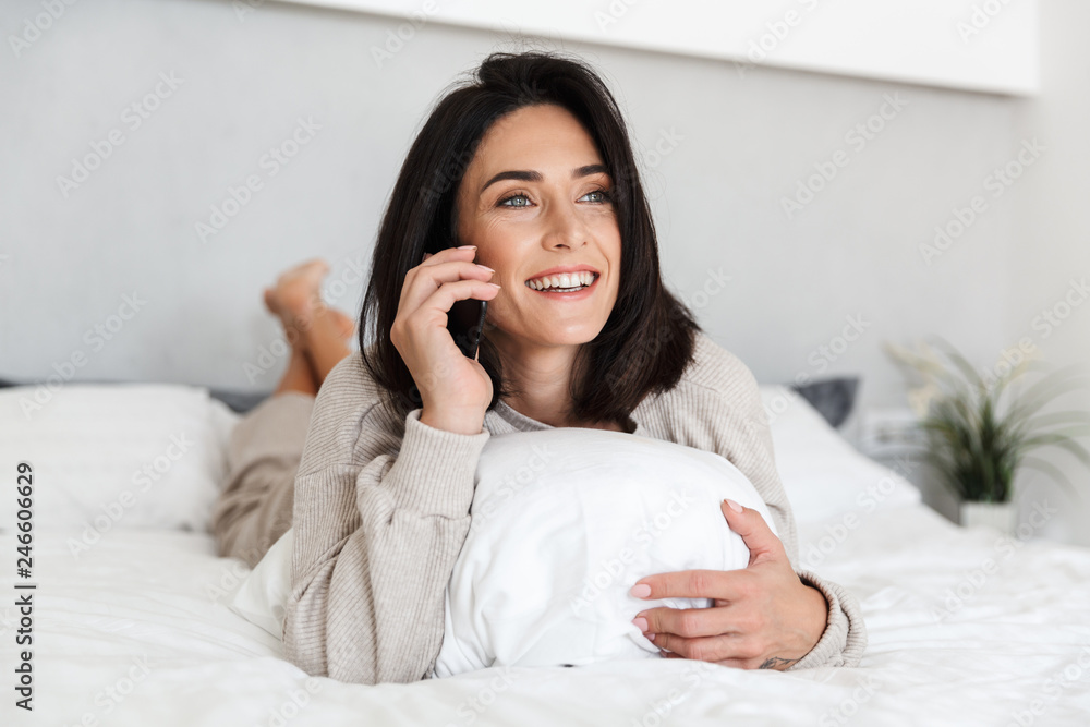 Photo of positive woman 30s using mobile phone, while lying in bed with white linen in bright room Photo of positive woman 30s using mobile phone, while lying in bed with white linen in bright room